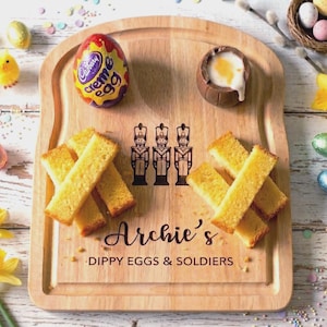 May include: A wooden serving board with the text "Archie's Dippy Eggs & Soldiers" and a toy soldier graphic. It features toast soldiers, a Cadbury Creme Egg, and a chocolate egg with a runny yolk. Easter theme.