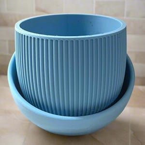 Op de afbeelding: Een lichtblauwe plantenpot met een geribbeld ontwerp en een bijpassende schotel. De pot is gemaakt van glad, mat materiaal.