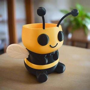May include: A yellow and black bee-shaped planter. The planter has a smiling face, black eyes, and antennae. The bee has a white wing on one side and black stripes. The planter is designed to hold small plants or succulents.