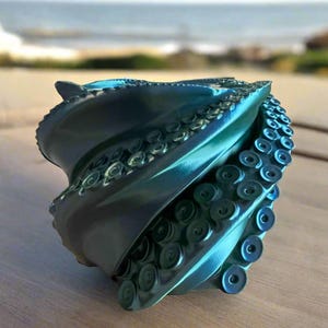 May include: A teal-colored, sculptural object resembling an octopus. The object has a spiral design with multiple tentacles featuring suction cups. The sculpture is on a wooden surface with a blurred background of the ocean and sky.