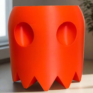 Puede incluir: Maceta de cerámica naranja con forma de fantasma con dos agujeros redondos para los ojos. La maceta tiene un borde inferior dentado.