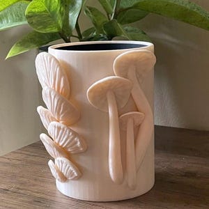 Puede incluir: Una maceta cilíndrica de cerámica blanquecina con un borde interior negro. La maceta está adornada con diseños detallados de hongos y hojas de color rosa claro. La maceta está sobre una superficie de madera, con plantas verdes en el fondo.