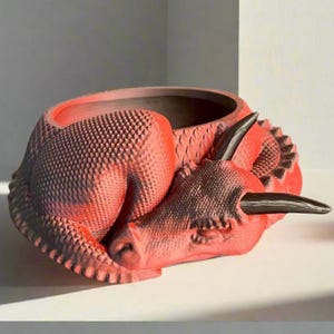 Puede incluir: Un macetero de cerámica roja en forma de dinosaurio con una superficie texturizada. El dinosaurio está durmiendo con la cabeza apoyada en su cuerpo. El macetero tiene un cuerno negro y un ojo negro.