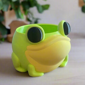 Może przedstawiać: Jasnozielona doniczka w kształcie żaby z żółtą paszczą i dużymi czarnymi oczami. Doniczka wykonana jest z gładkiego materiału i ma okrągły otwór. Żaba ma małe stopy i stoi na jasnej powierzchni.
