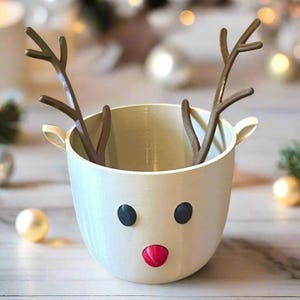 May include: A cream-colored reindeer-themed container with brown antlers, black eyes, and a red nose. The container has two small handles and is likely used for holiday decorations or as a festive gift.