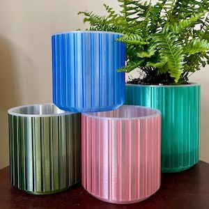 May include: Four cylindrical plant pots in blue, green, and pink. Each pot has vertical ridged detailing. One pot contains a green plant. The pots are arranged on a dark wooden surface.