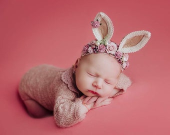 May include: A sleeping baby wearing a pink knitted onesie and a headband with pink flowers and bunny ears. The baby is lying on a pink background.