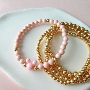 Peut inclure: Un ensemble de quatre bracelets, deux roses et deux dorés. Les bracelets roses ont une finition mate et les bracelets dorés ont une finition brillante. L'un des bracelets roses a un charme tête de Mickey Mouse rose.