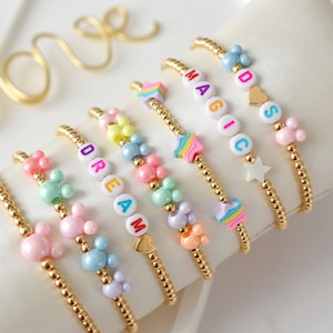 Könnte beinhalten: Ein Set aus acht goldenen Perlenarmbändern mit bunten Buchstabenperlen, die die Wörter "DREAM", "MAGIC" und "DADS" buchstabieren. Jedes Armband hat einen Anhänger, darunter ein Herz, ein Stern und ein Mickey-Maus-Kopf.