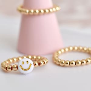 May include: A gold beaded ring with a white smiley face charm. The ring is on a pink display stand.
