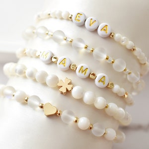 Op de afbeelding: Vijf witte kralenarmbanden met gouden accenten. De armbanden spellen "EVA" en "MAMA" met een gouden hart en een klavertje vier bedel.