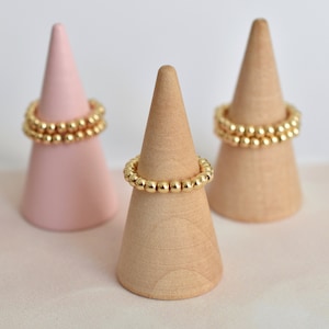 Könnte beinhalten: Drei goldene Perlenringe, die auf kegelförmigen Ringhaltern aus Holz und rosa präsentiert werden.