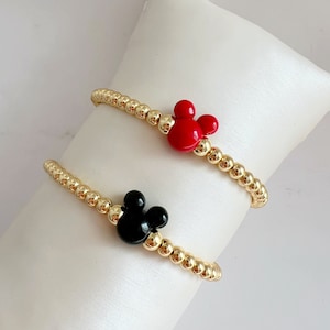 Peut inclure: Deux bracelets en perles dorées avec un charme tête de Mickey Mouse noir et rouge. Les bracelets sont sur un coussin blanc.