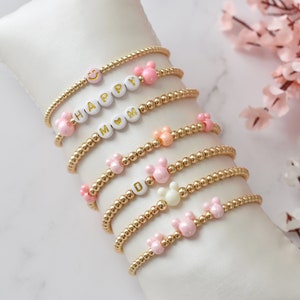 Peut inclure: Un ensemble de sept bracelets en perles dorées avec des perles roses et blanches. Les bracelets présentent différents charmes, dont un smiley, le mot "HAPPY", le mot "MOM" avec un cœur et des oreilles de Mickey Mouse.
