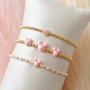 Peut inclure: Trois bracelets en perles dorées avec des breloques roses. Le bracelet du haut a un charme smiley, le bracelet du milieu a deux breloques en forme d'oreilles de souris, et le bracelet du bas a une breloque en forme d'oreille de souris.
