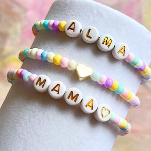 Peut inclure: Deux bracelets en perles colorées avec des perles de lettres blanches qui épellent les noms "ALMA" et "MAMA" avec un charme de cœur en or.