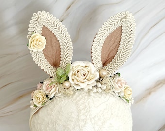 May include: A white lace headband with a floral design and bunny ears. The headband is decorated with white and pink flowers, green leaves, and pearl-like beads.
