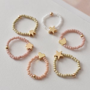 May include: Six beaded rings with gold charms. The charms include a crown, a flower, a heart, a star, and two different star shapes.