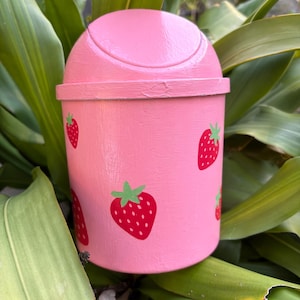May include: A pink, small trash can with a lid, decorated with red strawberries and green leaves. The trash can is made of wood and has a smooth, painted finish. The strawberries are evenly spaced around the can.