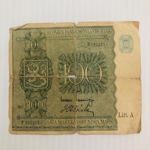May include: A vintage 100 Markka banknote from Finland. The note is green and features the Finnish coat of arms. The text "SUOMEN PANKKI FINLANDS BANK" is printed at the top of the note. The note is torn and worn.