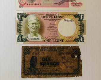 Könnte beinhalten: Eine Sammlung von fünf alten Banknoten aus verschiedenen afrikanischen Ländern. Die Banknoten zeigen Porträts historischer Persönlichkeiten, darunter ein Mann in Militäruniform, ein Mann mit traditionellem Hut und ein Mann im Anzug. Die Banknoten sind in unterschiedlichem Zustand, einige zeigen Anzeichen von Verblassen und Verfärbung.