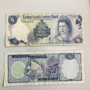 May include: A vintage Cayman Islands one dollar bill featuring a portrait of Queen Elizabeth II and a depiction of a ship. The bill is printed on paper with a light brown background and has a blue and white design. The text "Cayman Islands Currency Board" and "One Dollar" is printed on the bill.
