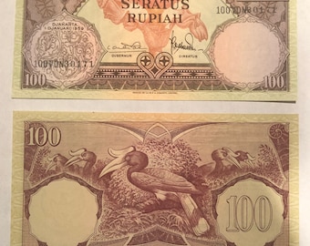Op de afbeelding: Twee Indonesische 100 Rupiah bankbiljetten. De voorkant toont de tekst "BANK INDONESIA" en "SERATUS RUPIAH", met een bloemmotief. De achterkant toont neushoornvogels en het nummer 100. De bankbiljetten zijn voornamelijk bruin en beige.