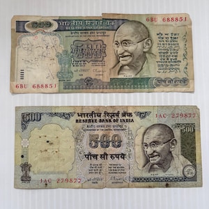 Puede incluir: Dos billetes indios de 500 rupias. Los billetes presentan un retrato de Mahatma Gandhi y texto en varios idiomas. El billete superior tiene el número de serie 6BU 68885J. El billete inferior tiene el número de serie JAC 279877.