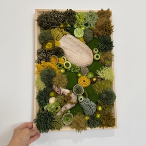 Puede incluir: Una obra de arte mural rectangular con marco de madera que presenta un diseño texturizado y tridimensional. La obra de arte incorpora varios tonos de verde, amarillo y marrón, con una piedra oblonga de color claro y un trozo de madera flotante.