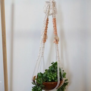 May include: A white macrame plant hanger with peach accents holds a wooden bowl filled with green foliage. The hanger is suspended from a ceiling hook.