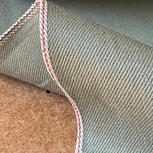 May include: A piece of olive green selvedge denim fabric with a distinct diagonal twill weave. The edge of the fabric is folded over to reveal a white selvedge ID with a thin, vibrant red line running vertically along the finished edge.