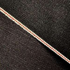 May include: Close-up of two different types of denim fabric. One is a dark indigo denim with a subtle twill weave. The other is a black denim with a more pronounced twill weave. A white and red stitching detail separates the two fabrics.