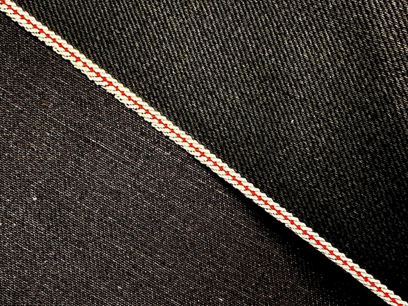 May include: Close-up of two different types of denim fabric. One is a dark indigo denim with a subtle twill weave. The other is a black denim with a more pronounced twill weave. A white and red stitching detail separates the two fabrics.