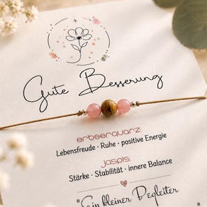 Könnte beinhalten: Ein zartes Armband mit zwei durchscheinenden rosa Perlen und einer zentralen braun gestreiften Perle an einer dünnen beigen Kordel, befestigt auf einer weißen Karte mit dem Text "Gute Besserung," "erbeerquarz," "jaspis," und "Ein kleiner Begleiter auf dem Weg zur Besserung."