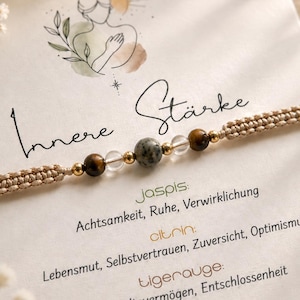 Könnte beinhalten: Ein beiges, geflochtenes Armband mit einer zentralen grauen Jaspisperle, flankiert von zwei klaren Quarzperlen, zwei braunen Tigeraugenperlen und kleinen goldfarbenen Metallzwischenperlen. Das Armband liegt auf einer Karte mit dem Text Innere Stärke.