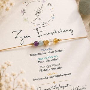 Könnte beinhalten: Ein feines Armband mit goldfarbenem Band, einem zentralen Herzanhänger aus Metall sowie violetten, hellblauen, klaren und gelben Perlen, befestigt auf einer weißen Karte mit deutschem Text und floralen Strichzeichnungen.