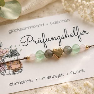 Könnte beinhalten: Ein Perlenarmband auf einer Karte mit dem Text 'glücksarmband • talisman', 'Prüfungshelfer' und 'labradorit • amethyst • fluorit'. Das Armband hat ein zentrales goldfarbenes Herz-Element, flankiert von grauen, grünen und rosafarbenen Perlen.