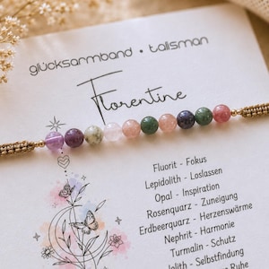 Peut inclure: Un bracelet composé de dix perles en pierres gemmes multicolores sur un cordon tressé marron, présenté sur une carte blanche avec le texte "glücksarmband • talisman", "Florentine" et une liste de propriétés incluant "Fluorit - Fokus".