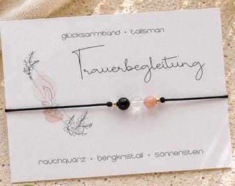Könnte beinhalten: Ein schwarzes Armband aus Kordel mit drei Perlen: ein schwarzer Rauchquarz, ein klarer Bergkristall und ein pfirsichfarbener Sonnenstein, getrennt durch kleine goldfarbene Zwischenelemente. Das Armband ist auf einer weißen Karte mit dem Text Trauerbegleitung befestigt.