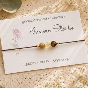 Könnte beinhalten: Ein braunes Armband aus Kordel mit drei Perlen: ein gesprenkelter beiger Jaspis, ein durchsichtiger gelber Citrin und ein braunes Tigerauge, präsentiert auf einer weißen Karte mit Rosenillustration und dem Text glücksarmband • talisman Innere Stärke Jaspis • citrin • tigerauge.