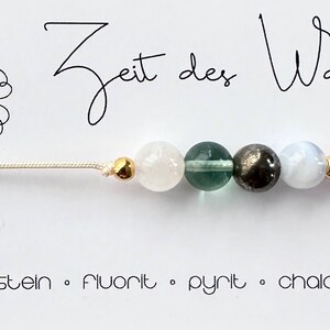May include: A delicate bracelet featuring four gemstone beads: moonstone, fluorite, pyrite, and chalcedony. The beads are strung on a beige cord, accented with gold-colored beads. The card reads "Zeit des Wandels" and lists the gemstones.