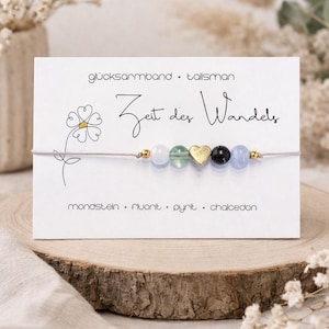 Può includere: Un braccialetto di perline con un ciondolo a forma di cuore dorato e varie perline di pietre preziose, tra cui pietra di luna, fluorite e calcedonio. Il braccialetto è esposto su una superficie di legno con una carta che recita "Zeit des Wandels".