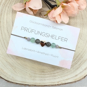 Könnte beinhalten: Ein Armband mit herzförmigem Anhänger, präsentiert auf einer Karte mit deutschem Text. Das Armband hat grüne, lila und graue Perlen. Auf der Karte stehen auch die Worte "Labradorit, Amethyst, Fluorite."