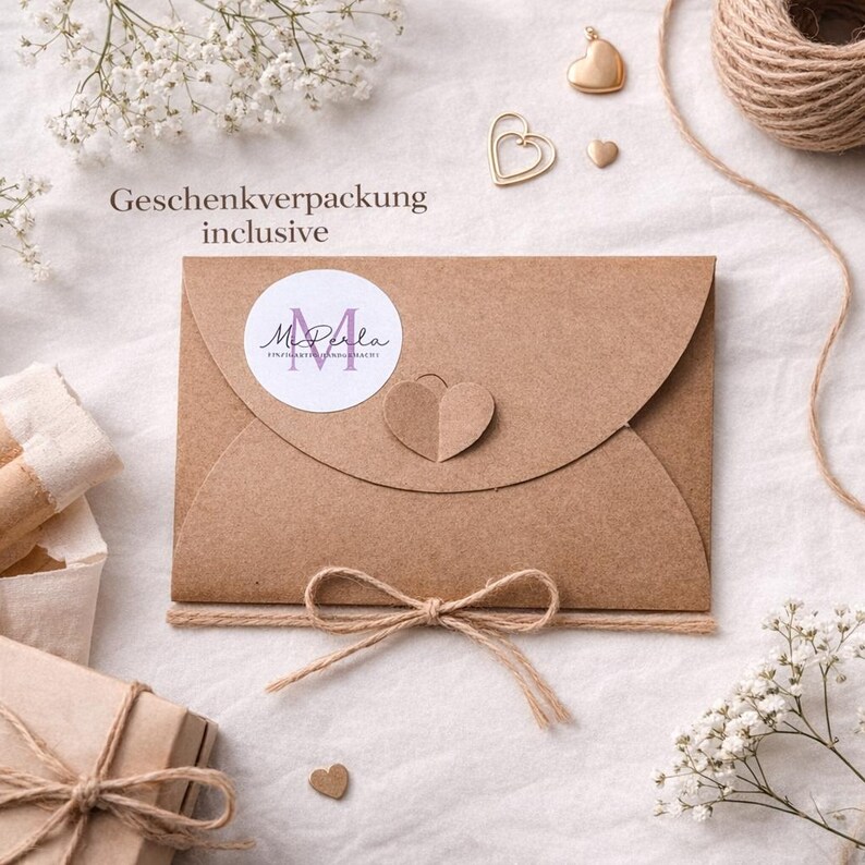 May include: A brown paper envelope with a heart-shaped cutout and a twine bow. The envelope has a circular sticker with the text "M. Perla". The words "Geschenkverpackung inclusive" are printed above the envelope. Small gold heart-shaped charms and twine are also visible.