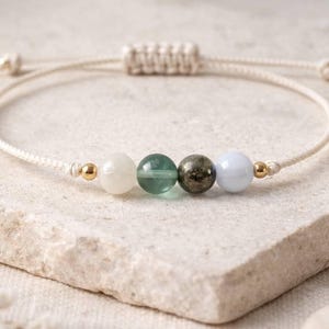 May include: A beaded bracelet featuring a cream-colored cord with four gemstone beads: white, green, dark gray, and light blue. Gold-colored beads accent the design. The bracelet is adjustable.