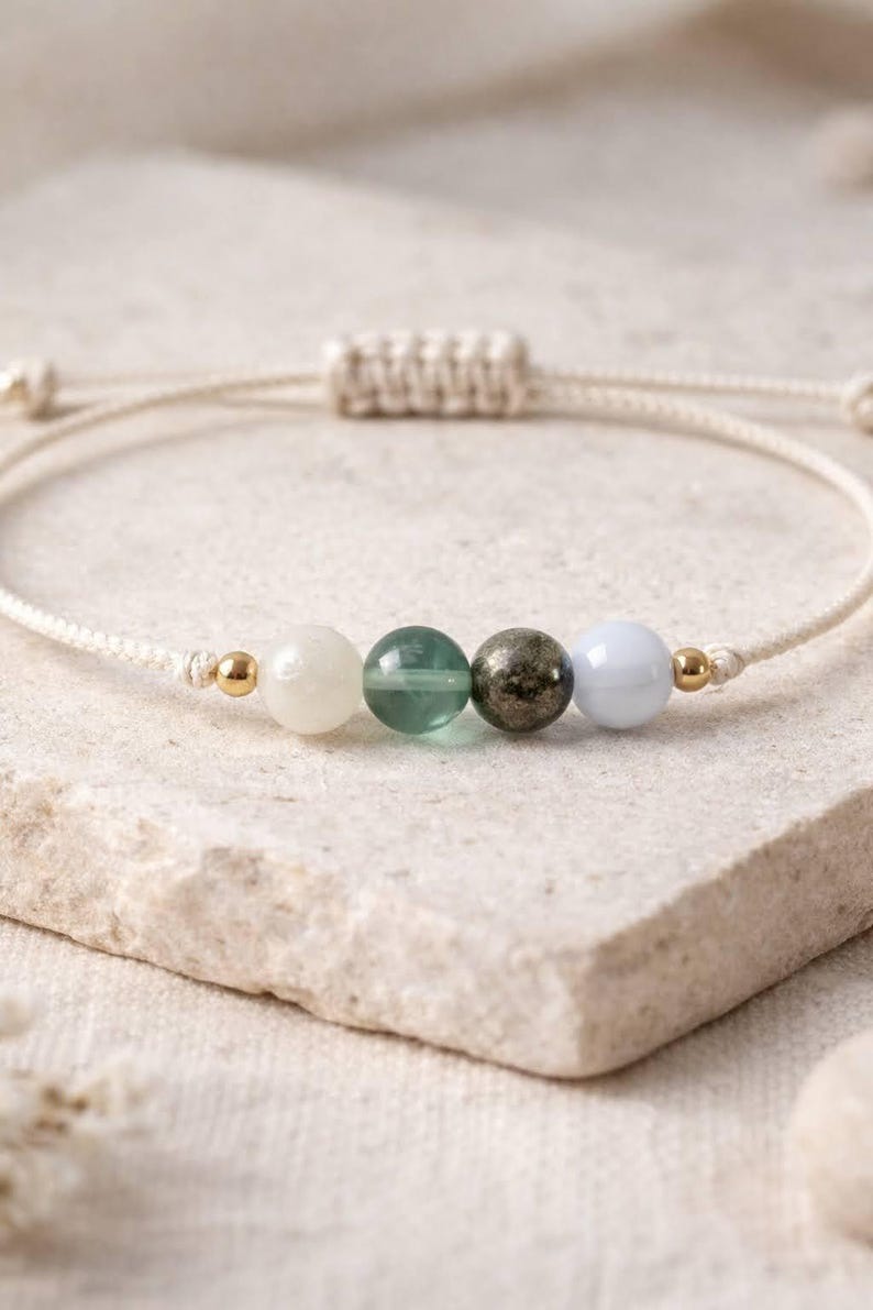 May include: A beaded bracelet featuring a cream-colored cord with four gemstone beads: white, green, dark gray, and light blue. Gold-colored beads accent the design. The bracelet is adjustable.