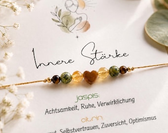 May include: A delicate bracelet featuring a central brown heart-shaped bead flanked by faceted yellow beads, green speckled beads, and small brown tiger eye beads on a thin gold-colored cord, displayed on a card with the text Innere Stärke.