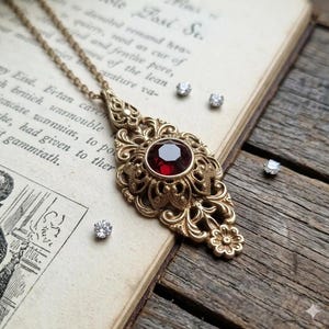 May include: A gold-toned metal pendant necklace featuring an intricate filigree design and a central round faceted deep red gemstone. The pendant is suspended from a matching gold-toned chain, accompanied by four small, clear, round faceted stones.