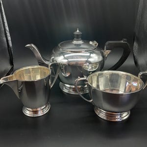 Puede incluir: Un juego de té plateado que incluye una tetera, una jarra para la crema y un azucarero. La tetera tiene un asa negra y un adorno decorativo. El juego está hecho de metal pulido y se encuentra sobre una superficie negra. La jarra para la crema tiene un pico.