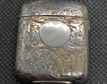 Könnte beinhalten: Ein silbernes Streichholzsafe mit einem verzierten gravierten Design. Das Streichholzsafe hat einen Klappdeckel und eine runde Öffnung auf der Vorderseite.